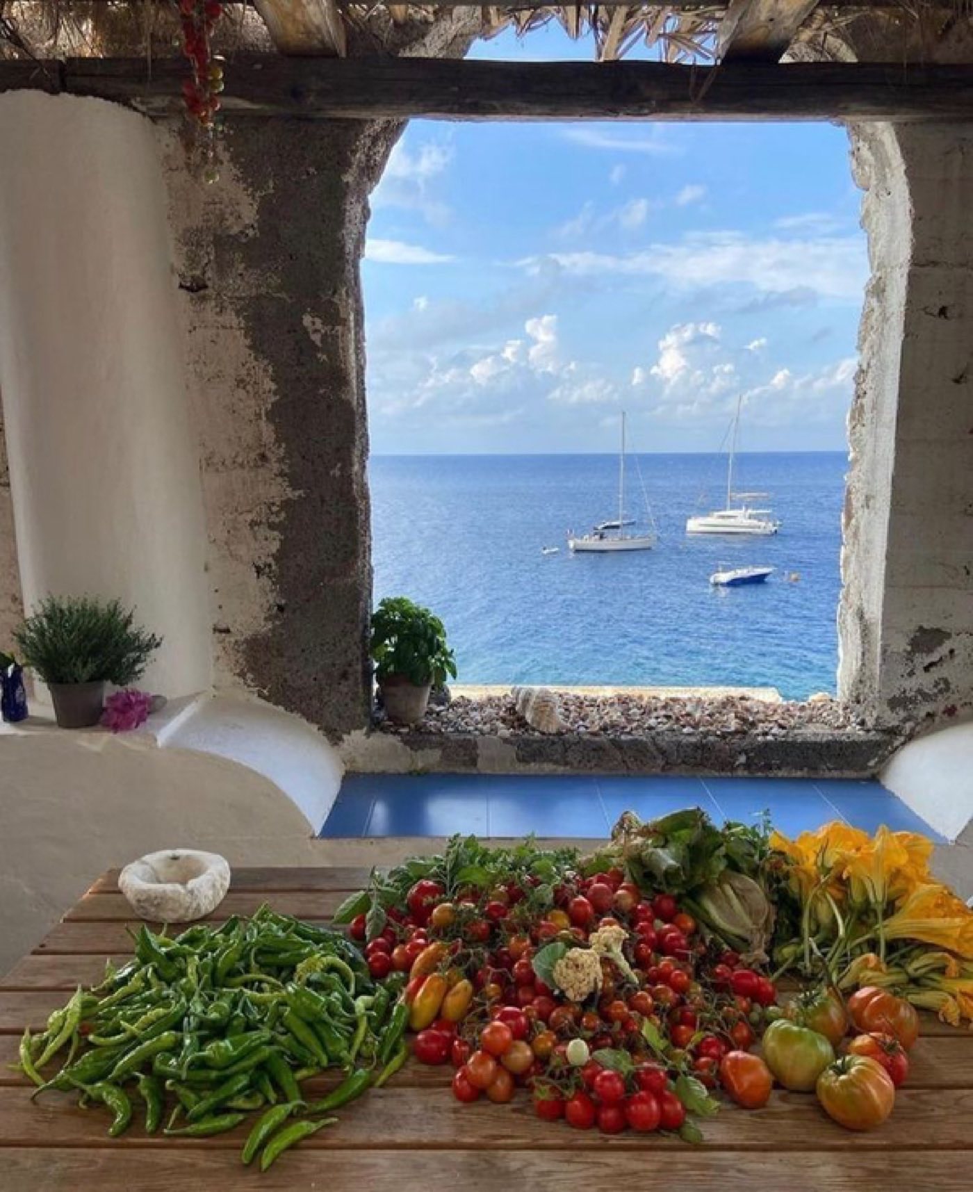 A stone window overlooking the Mediterranean sea, with freshly harvested peppers, tomatoes, and squash blossoms laid out on a wooden table